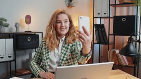 Hello. Happy businesswoman waves hand hi gesture greeting welcomes someone on smartphone, online selfie webinar, remote distance conversation. Freelance sitting at desk using laptop in home office