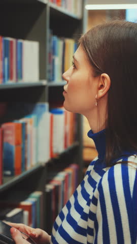 Vertical screen: Young woman in blue and white striped sweater selecting book from library shelf, holding tablet. Education and resource searching concept
