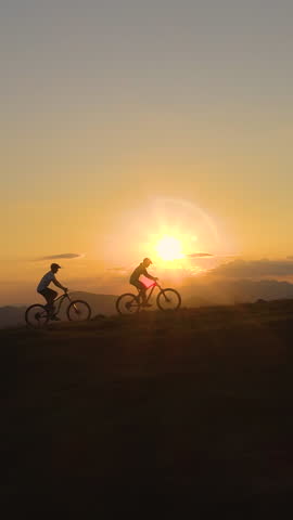 AERIAL, SILHOUETTE, LENS FLARE: Breathtaking evening sunbeams shine on the tourists riding ebikes in the scenic nature. Three downhill bikers pedalling their bicycles up along grassy hill at sunrise.