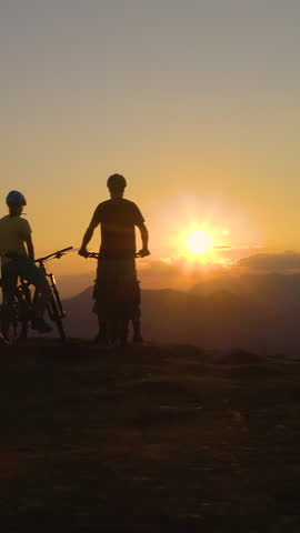 DRONE, SILHOUETTE, LENS FLARE: Mountain bikers stop and observe the sunset after an adrenaline filled downhill adventure. Three friends watch the sunset after a bicycle ride in the scenic mountains Royalty-Free Stock Footage #3446065619