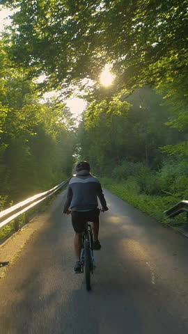 SUN FLARE, DRONE: Flying behind unrecognizable man riding his bicycle at sunrise along empty road leading through the beautiful sunlit forest. Athletic male enjoying a relaxing bike ride at sunset.