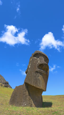 LOW ANGLE, VERTICAL: Scenic shot of a black rock formation formed into a human face set in the middle of a grassy hill on Easter Island. White clouds drift over the large moai sculpture on a sunny day