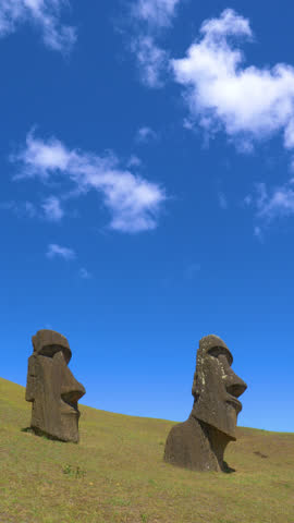 VERTICAL: White clouds drift above two big moai statues on remote exotic island in the Pacific. Cool shot of statues made of volcanic matter erected in the middle of a grassy hill on Easter Island