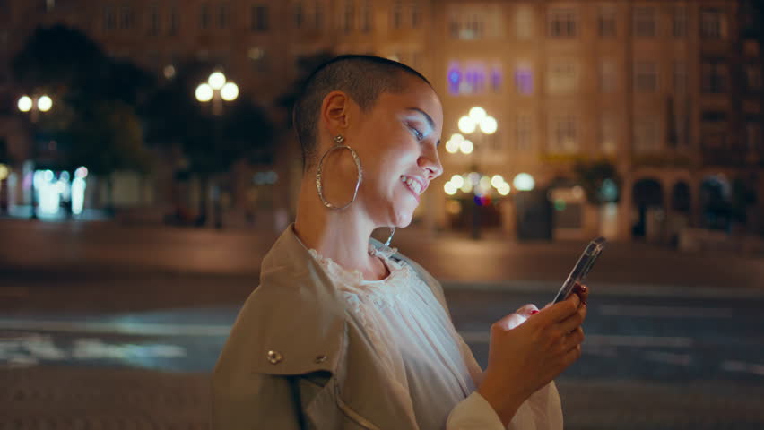 Cheerful lady scrolling cellphone at night city closeup. Happy short hair woman texting cellphone waiting meeting on evening street. Trendy informal girl watching social media standing dark urban area - Powered by Shutterstock - Get 15% off with code: PIKWIZARD15