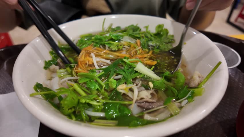 Asian woman eating Vietnamese soup dish Pho that consisting of broth, rice noodles, herbs, and beef pork chicken meat with scallion, onion, cilantro (coriander leaves), bean sprouts, lime, chili oil.