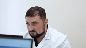 Middle aged doctor with in clinic. Clip. Bearded man in white coat looking at computer monitor.