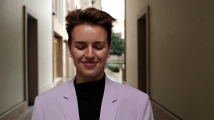 Young woman listening to music on headphones and dancing. She has short hair and she is under a corridor. She is wearing a purple blazer.