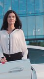 A beautiful dark-haired business woman in a white shirt is standing near a white car. The woman poses for the camera, behind her is a glass business center.Vertical video.