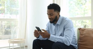 Positive handsome young African man talking on video call, enjoying online business conference chat on mobile phone, sitting on home sofa, speaking at screen, smiling