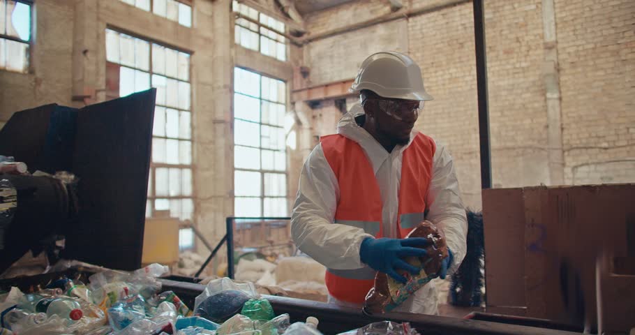 A man with Black skin in a white uniform and an orange vest in protective blue gloves sorts through garbage and plastic bottles depending on color at the Large Waste Recycling Plant Royalty-Free Stock Footage #3446386277