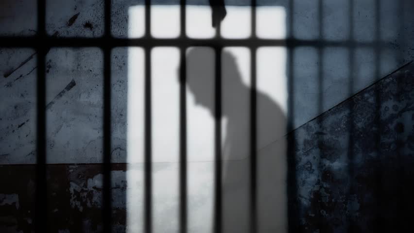 Silhouette: a desperate man, hands pressed against his face, his back bowed under the weight of troubles, confined behind shadowed prison bars, his image cast upon a weathered wall.
 Royalty-Free Stock Footage #3446442663