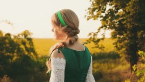 Young beautiful beauty goes sunflower field on backdrop summer grass trees. Medieval green dress costume white blouse Smiling face. Hairstyle two blond braids back rear view. Princess enjoy nature 