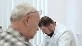 Male doctor working at the table, writing prescription. Clip. Old male patient at the hospital.