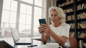 Senior woman freelancer work on laptop. Old lady receiving message in smartphone reading typing answer sitting at table at home. Freelance distant remote job, work. Home office pensioner, retirement. - Powered by Shutterstock - Get 15% off with code: PIKWIZARD15