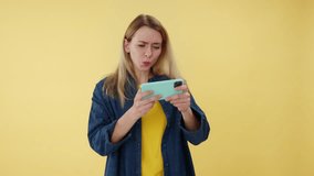 Energetic lady gamer holding personal smartphone horizontally and playing online video game indoors. Active caucasian female using modern gadget for fun time. Isolated over yellow studio background.