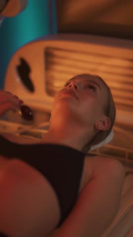 Sun treatments in a horizontal solarium, young woman takes a sun bath in a solarium, uses eye protection, warm light, vertical video for social networks.