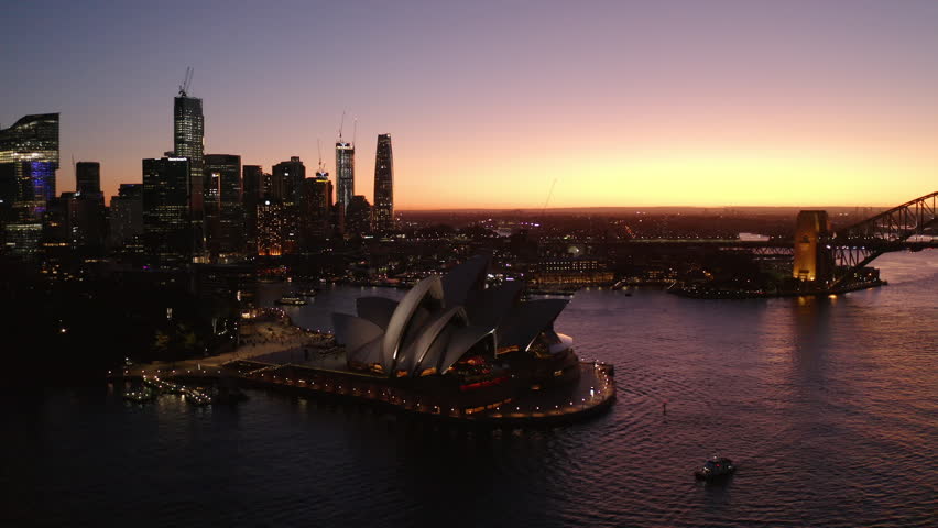 Aerial Panning Shot Of Illuminated Sydney Opera House In Modern City Against Orange Clear Sky, Drone Flying Over Tasman Sea