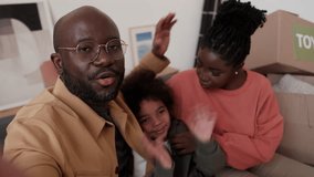 POV of happy African American parents and little kid looking at camera, waving and chatting via online video call on couch with unpacked boxes after moving in new home
