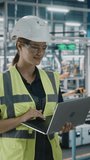 Vertical Portrait of Female Automotive Industry 4.0 Engineer in Safety Uniform Using Laptop at Car Factory Facility. Happy Assembly Plant Specialist Working on Manufacturing Modern Electric Vehicles.