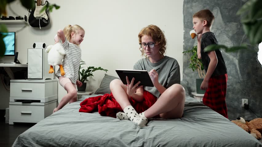 A mother woman works from home, and uses a tablet to communicate with colleagues. Children run, mess around and make loud noises at home. Royalty-Free Stock Footage #3447115755