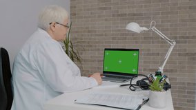 Woman doctor watches on laptop with green screen. Nurse in uniform and lab coat clothes sitting in hospital office room using laptop.