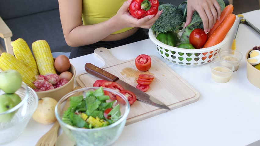 A young woman with a figure is picking vegetables and cutting them, making a salad in the kitchen at home. Happy woman is preparing a fresh healthy salad with many vegetables in the kitchen.