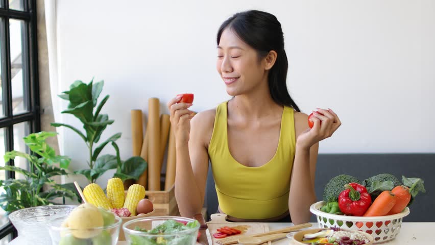 Beautiful healthy Asian woman eating fresh tomatoes in the kitchen at home. Happy beautiful female healthy influencer eating fresh tomatoes.