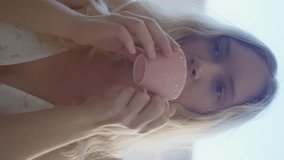 A young girl is delicately holding a pink cup with her thumb and gesturing towards her lips while she takes a sip. Her jaw is relaxed as she enjoys her drink Slow motion. Vertical video.