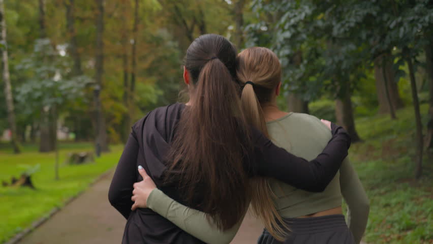 Back view two Caucasian girls active sportswomen happy ladies female friends walking together in park outdoors backside women laughing after sport training in city give high five good friendship hug