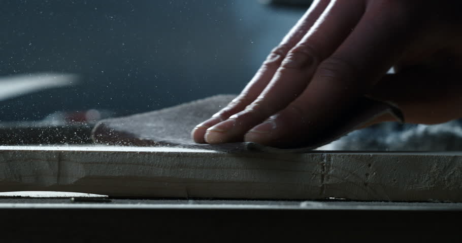 Super slow motion close up of artisan carpenter is sanding wood with hands and sand paper with flying wood sawdust on workbench in workshop at 1000 fps.