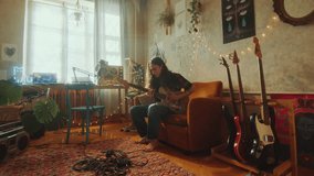 Young guy playing electric guitar while sitting on couch in a sunlit room with creatively designed interior, cozy string lights and retro tape recorders. Zoom shot - Powered by Shutterstock - Get 15% off with code: PIKWIZARD15