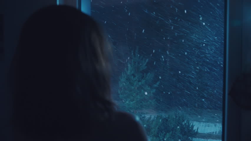 Anonymous girl standing by window looking at fabulous snowfall at home. Female person at blue night enjoying snow storm and snowflakes falling on green fir trees. Film grain pixel texture