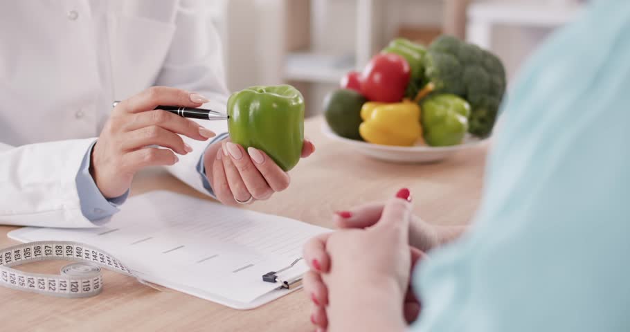 Crop professional dietician at table with vegetables, diet plan and tape measure holding green pepper in hand, giving dietary consultation to young woman, recommending healthy food and balanced menu Royalty-Free Stock Footage #3447359307