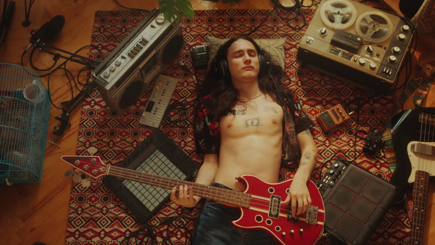 Young relaxed musician in headphones and open shirt playing electric guitar with eyes closed while lying down on floor among retro tape recorders and audio devices. Directly above view