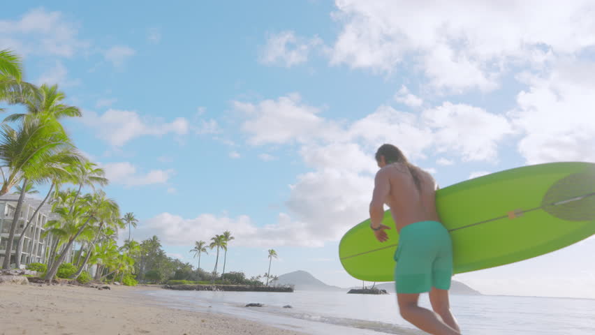 Young fit man with a surfboard running along the tropical beach island, under the cloudy sky, ready to conquer the waves and enjoy the thrill of surfing. Beautiful scenery with palm trees. Hawaii