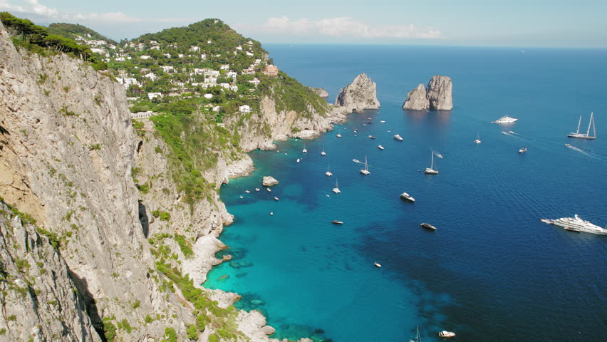 The iconic Faraglioni Sea Stacks emerge from Capri's azure sea. Towering rock formations, luxury vessels navigate the serene Mediterranean waters. Royalty-Free Stock Footage #3447454987