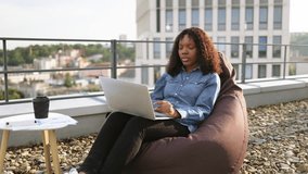 Business woman in casual denim shirt working on laptop. Professional businesswoman using digital device having video chat conference, sitting on beanbag on rooftop of modern building.