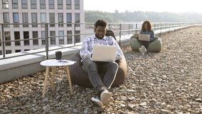 Delighted African American man screaming and raising hand up looking at laptop during coffee break on rooftop while his colleague working in blurred background. - Powered by Shutterstock - Get 15% off with code: PIKWIZARD15