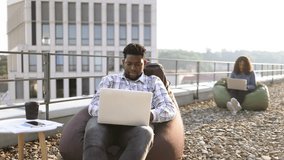 Delighted African American man screaming and raising hand up looking at laptop during coffee break on rooftop while his colleague working in blurred background. - Powered by Shutterstock - Get 15% off with code: PIKWIZARD15