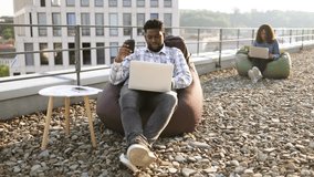 Charming African American man in casual shirt holding cup of hot drink while scrolling webpages on portable computer outdoors, while female colleague working in background. - Powered by Shutterstock - Get 15% off with code: PIKWIZARD15