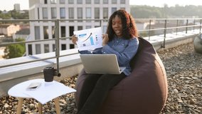 Young African female in casual clothes giving online presentation using headset and computer outside office. Remote manager brainstorming about statistics shown by infographics on terrace.