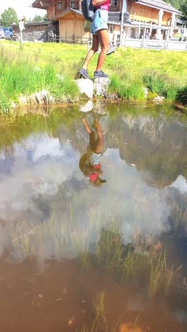 A woman in shorts stands by a pond, her reflection visible in the water. Surrounding her are plants, grass, trees, and the natural landscape of the Dolomites in Italy, Geisleralm Val Di Funes