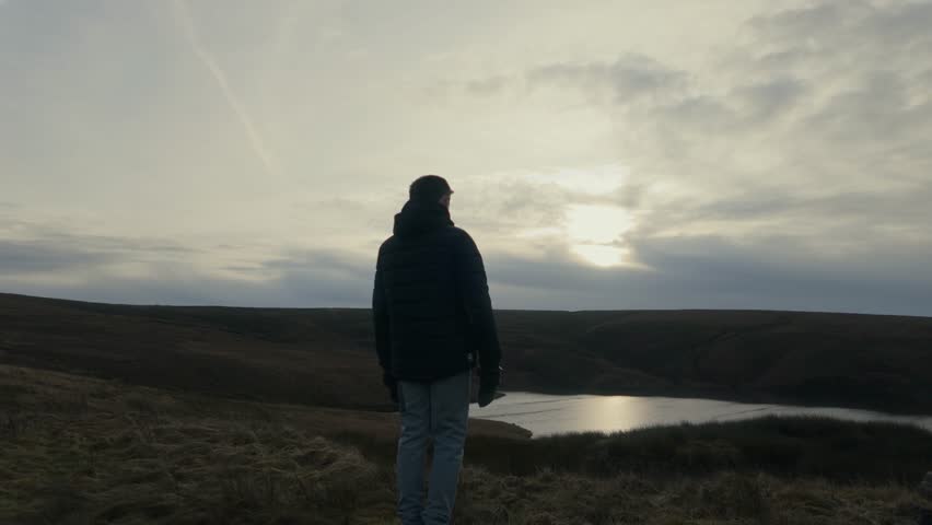 Young boy standing and looking out on to the winter landscape, with moorlands, fens, a lake and stormy winters sky. Sunset view of the Yorkshire landscape