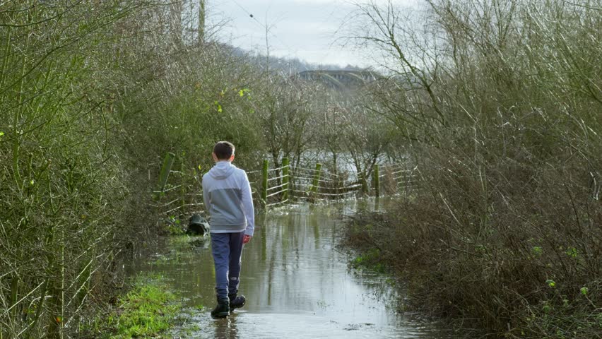 Young farmers boy walking along a flooded country path and wading through the flood lands. Global warming and extensive flooding in the winter of the UK. English cold spell.