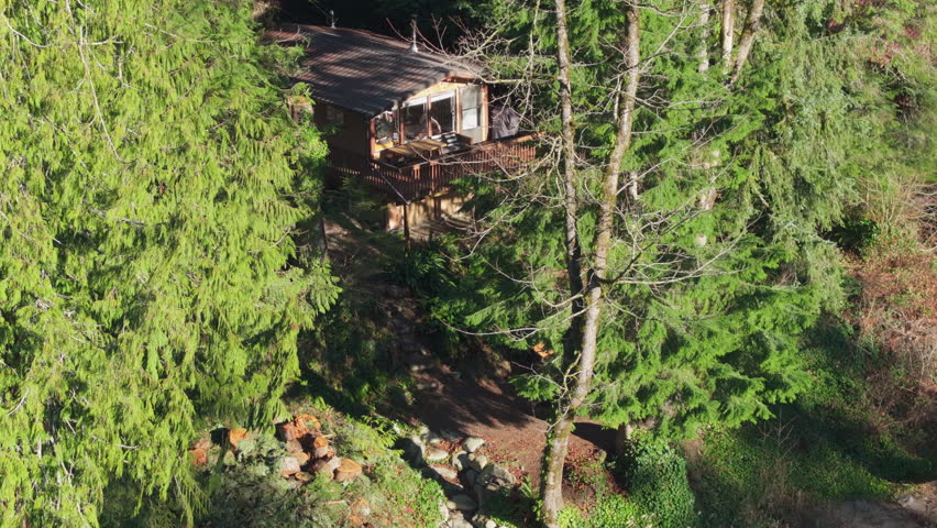 Small rustic countryside house hides between evergreen trees in lighted by golden sunshine. Mountain forest at blue river with Log cabin on sunny day. Two cabins in magical Washington at forest river