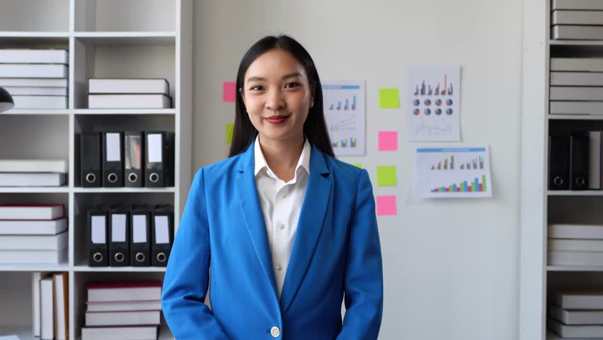 A charming Asian woman with a smile wearing a blue suit stands with her arms crossed confidently working at a start-up business within a company. Royalty-Free Stock Footage #3447621635