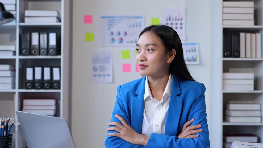 A charming Asian woman with a smile wearing a blue suit stands with her arms crossed confidently working at a start-up business within a company. Royalty-Free Stock Footage #3447621917