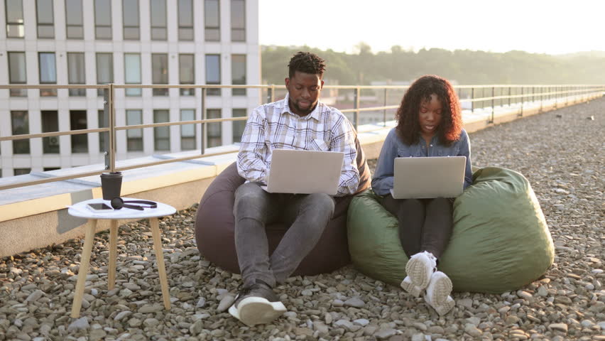 Beautiful young African American couple using laptops while sitting in beanbags at rooftop of office. Two business people working with modern devices sitting on rooftop terrace outside office. - Powered by Shutterstock - Get 15% off with code: PIKWIZARD15
