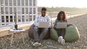 Beautiful young African American couple using laptops while sitting in beanbags at rooftop of office. Two business people working with modern devices sitting on rooftop terrace outside office. - Powered by Shutterstock - Get 15% off with code: PIKWIZARD15