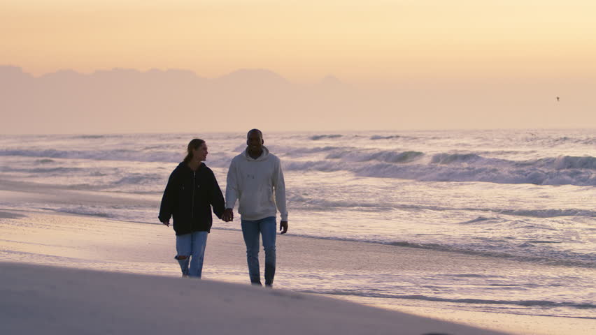 Full length shot of casually dressed loving young couple walking hand in hand along sandy shoreline watching beautiful sunrise morning over beach and sea in South Africa - shot in slow motion